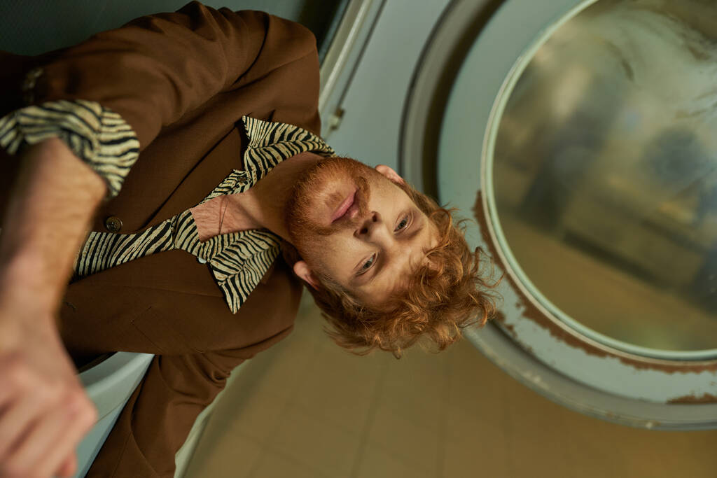 fashionable young redhead man in jacket posing in washing machine in public laundry - Photo, Image