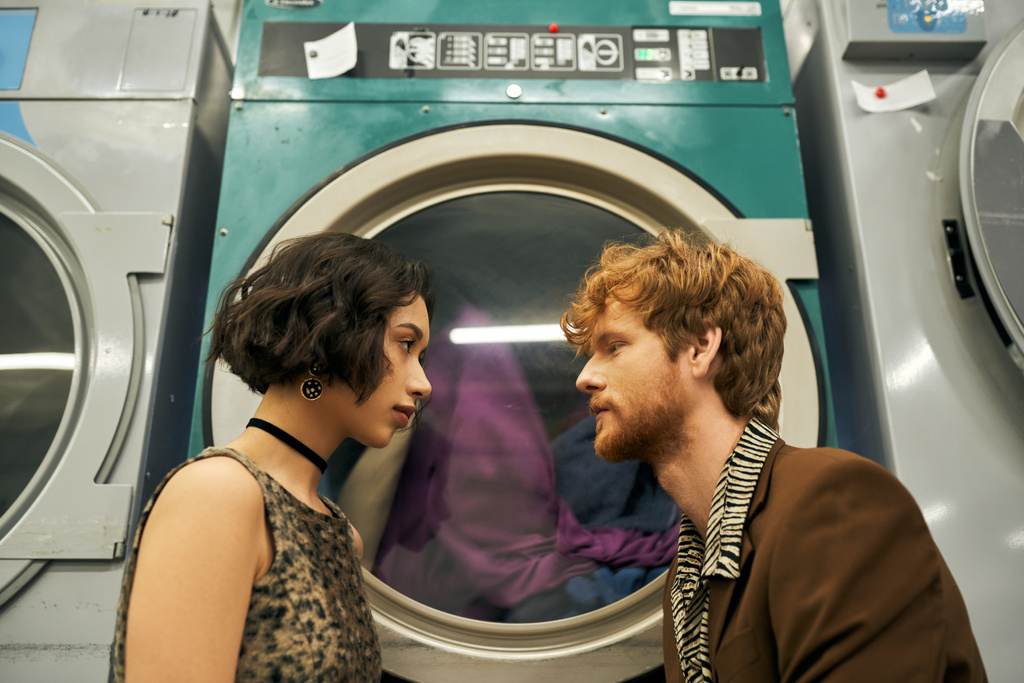 コインランドリーの洗濯機の近くにポーズするロマンチックでスタイリッシュな異人種間のカップルの側面 - 写真・画像