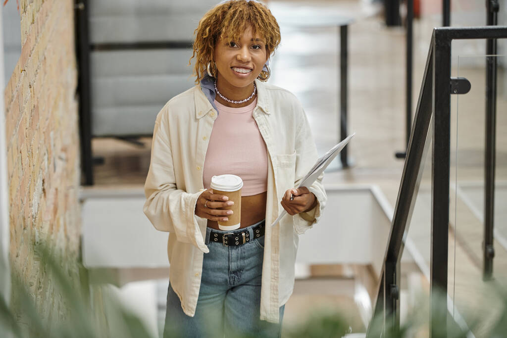 mujer afroamericana positiva con frenos sosteniendo taza de papel, café y documentos, proyecto de inicio - Foto, imagen