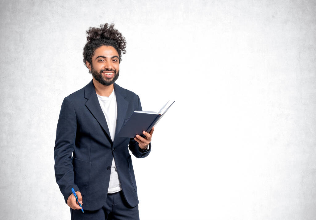 Lächelnder arabischer Geschäftsmann mit Notizbuch und Stift in der Hand, der vor leeren grauen Betonwänden in die Kamera blickt. Konzept der betriebswirtschaftlichen Ausbildung - Foto, Bild