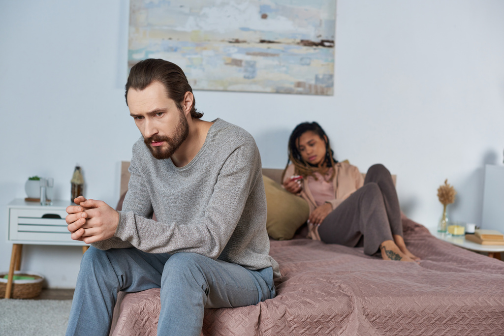 trauriger Mann sitzt im Bett, fühlt sich gestresst, afrikanisch-amerikanische Frau mit Schwangerschaftstest, Entscheidung - Foto, Bild