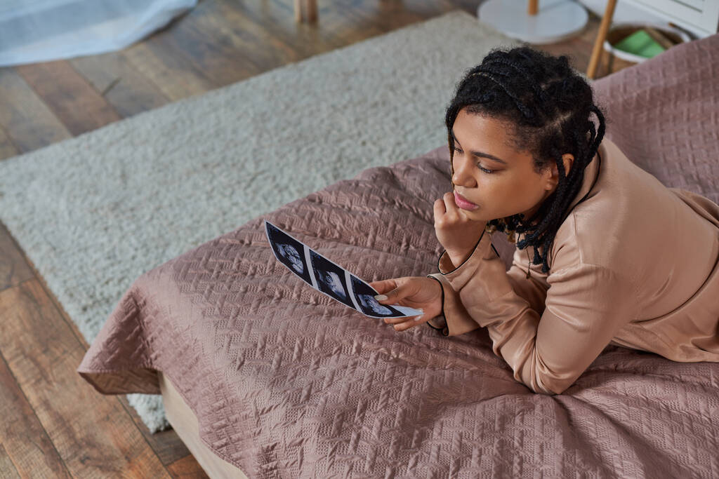 vista superior de la mujer afroamericana preocupada acostada en la cama, mirando ultrasonido, nonato, aborto - Foto, Imagen
