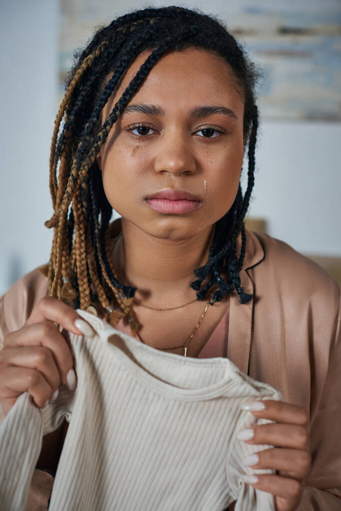 mujer afroamericana deprimida sosteniendo ropa de bebé y llorando en casa, concepto de aborto espontáneo - Foto, Imagen