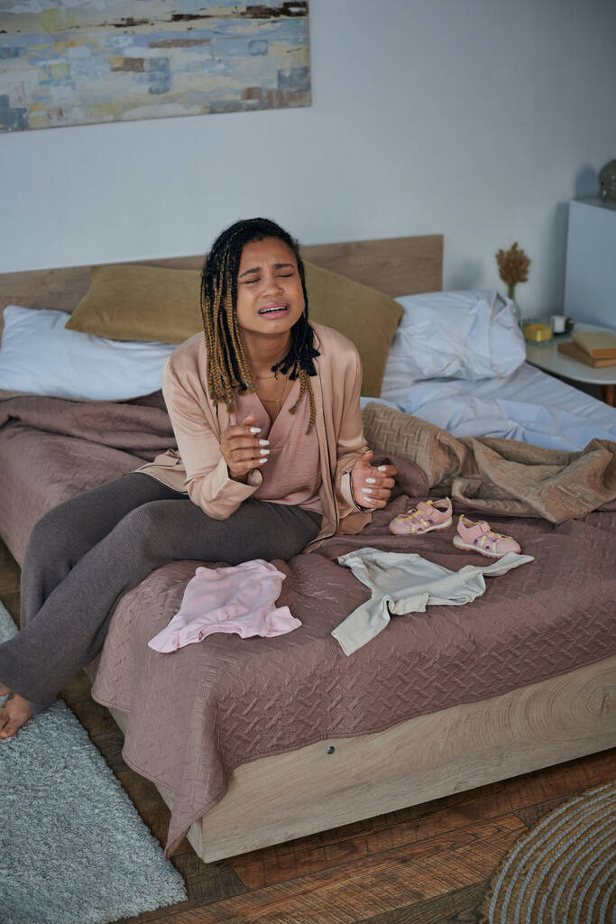 wanhoop, depressieve Afro-Amerikaanse vrouw huilt in de buurt van babykleertjes op bed, miskraam concept - Foto, afbeelding