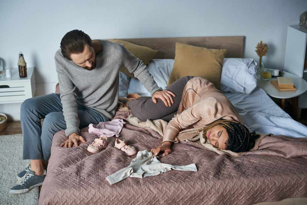 Trauer, Fehlgeburtenkonzept, Mann beruhigt afrikanisch-amerikanische Frau weint in der Nähe von Babykleidung, liegt auf dem Bett - Foto, Bild