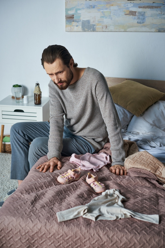 dolor, hombre deprimido llorando cerca de la ropa de bebé, sentado en la cama, concepto de aborto espontáneo, angustia - Foto, Imagen