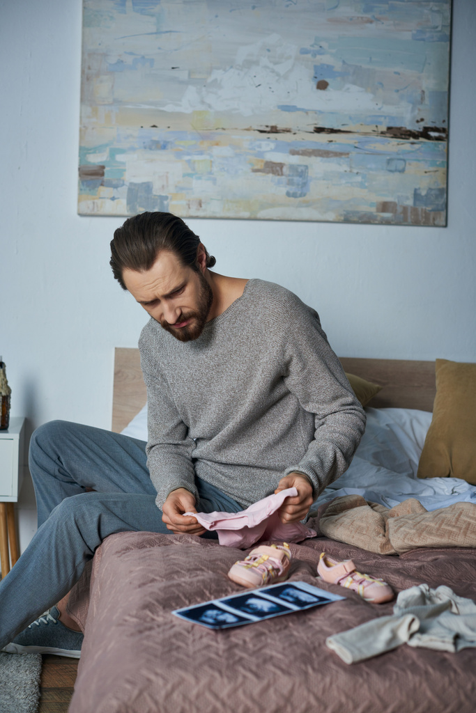 απώλεια, κατάθλιψη άνθρωπος κάθεται στο κρεβάτι κοντά στα ρούχα του μωρού και υπερηχογράφημα, αποβολή έννοια - Φωτογραφία, εικόνα