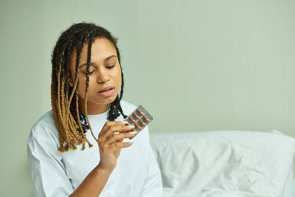 mujer afroamericana en bata de hospital sosteniendo pastillas, sala privada, aborto espontáneo, paciente en clínica - Foto, imagen