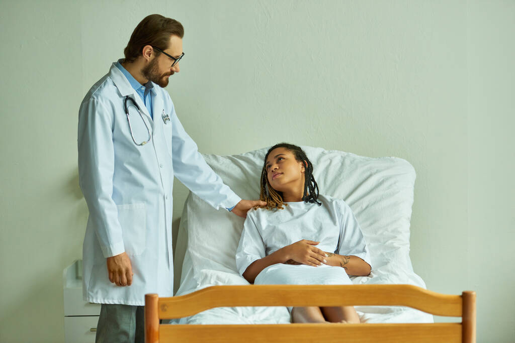 врач в очках, стоящий рядом с африканской американкой в больничном халате, в палате, пациент - Фото, изображение