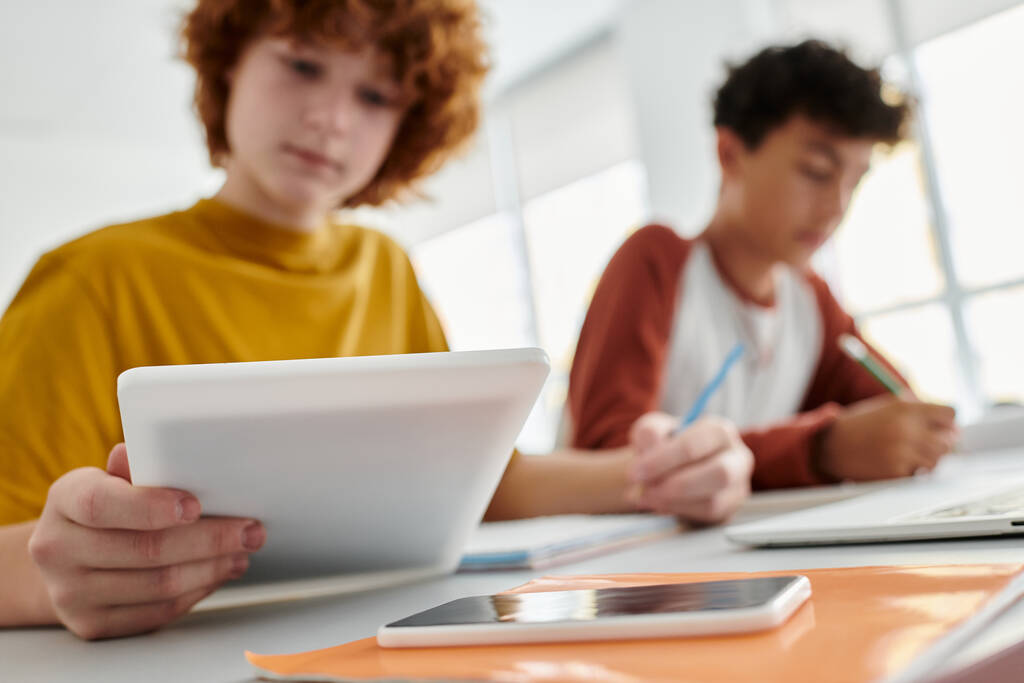 Escolar adolescente borrosa usando tableta digital cerca de amigo y portátil durante la lección en el aula - Foto, imagen
