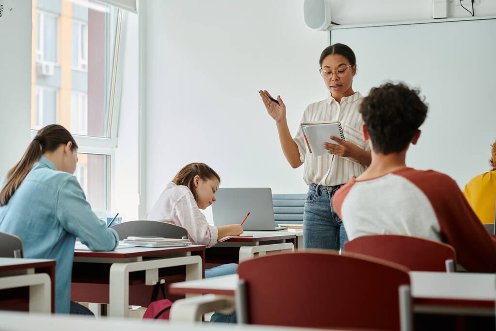 Professeur afro-américain avec ordinateur portable pointant et parlant pendant les cours avec des élèves adolescents en classe - Photo, image