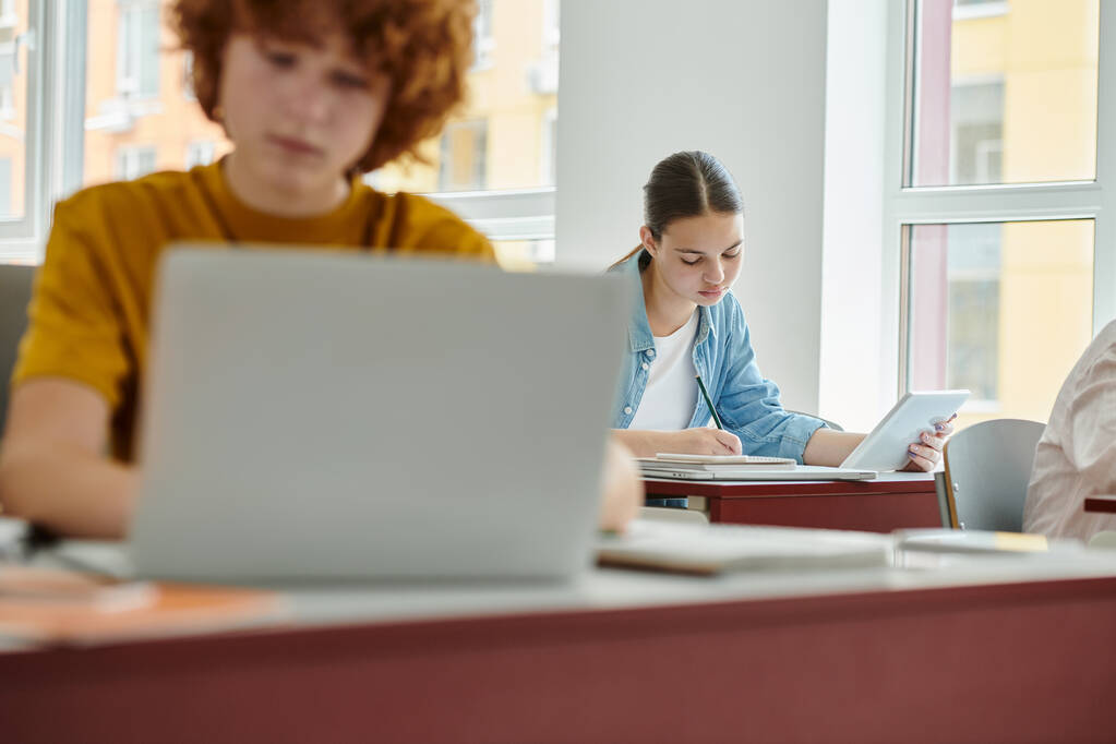 Adolescente colegiala sosteniendo tableta digital y la escritura en el cuaderno cerca borrosa compañero de clase en el aula - Foto, Imagen