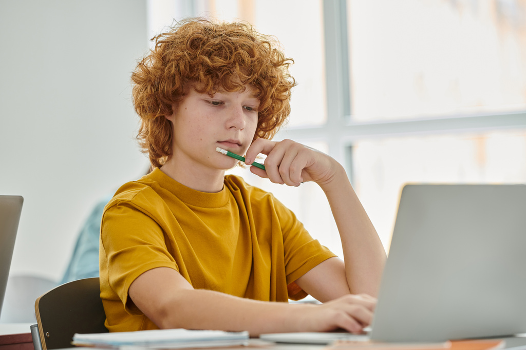 Kızıl saçlı liseli çocuk, elinde kalem tutuyordu ve sınıfta arka planda dizüstü bilgisayar kullanıyordu. - Fotoğraf, Görsel