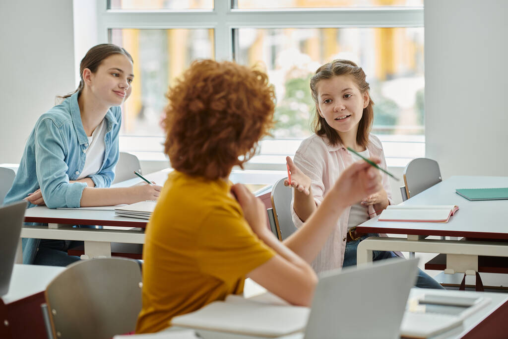 Positive teen schoolgirl pointing with hand while talking to classmate near devices in classroom - Photo, Image