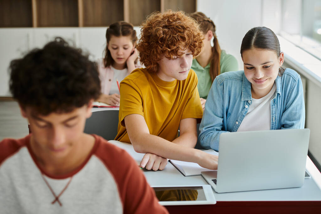 Kızıl saçlı liseli çocuk, bulanık okul dersinde gülümseyen sınıf arkadaşıyla birlikte dizüstü bilgisayar kullanıyor. - Fotoğraf, Görsel
