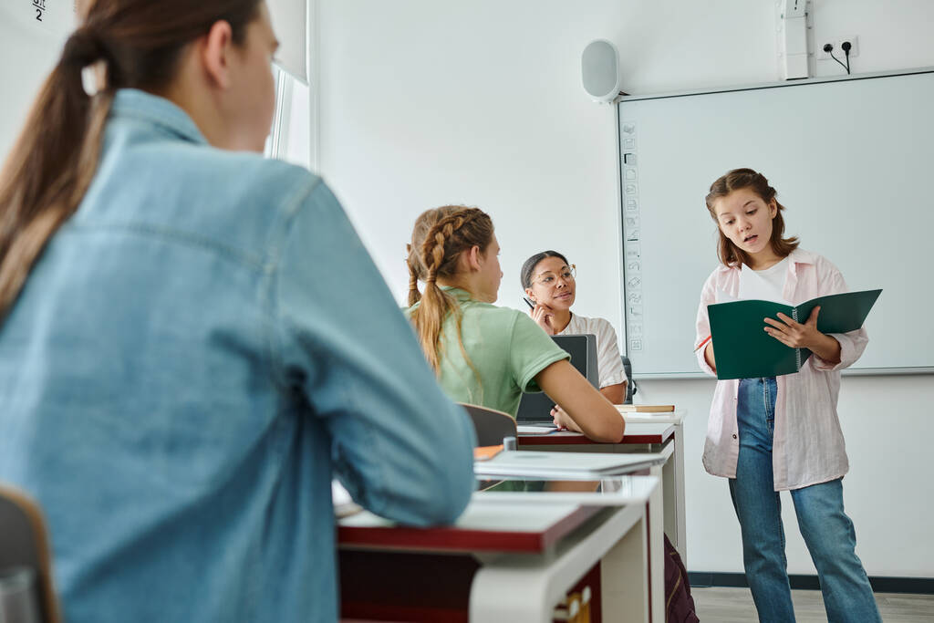 Schulmädchen spricht, während sie ihr Notizbuch in der Nähe eines lächelnden afrikanisch-amerikanischen Lehrers im Klassenzimmer hält - Foto, Bild