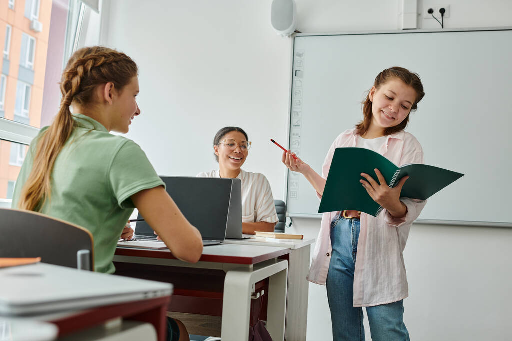 Usmívající se školačka mluví a drží notebook v blízkosti afrického amerického učitele a spolužáka ve třídě - Fotografie, Obrázek