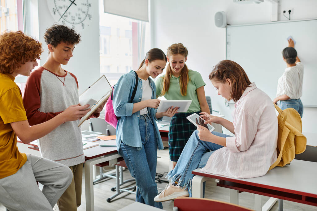 Alunos adolescentes alegres usando dispositivos perto de professor da américa africana em segundo plano em sala de aula - Foto, Imagem