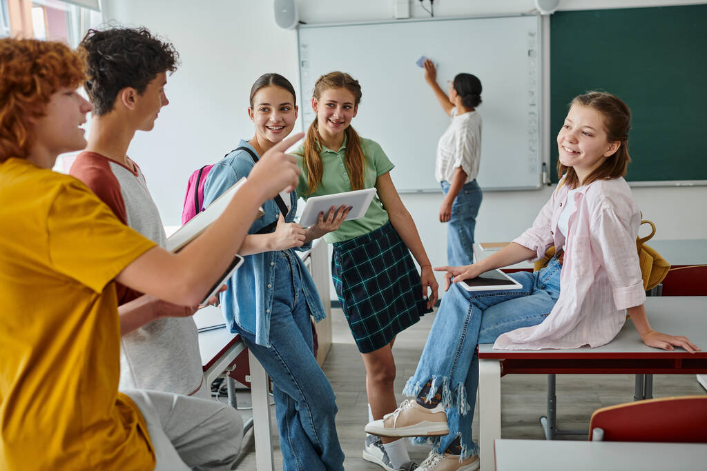 Estudantes sorridentes conversando e apontando com o dedo perto de alunas com dispositivos em sala de aula - Foto, Imagem