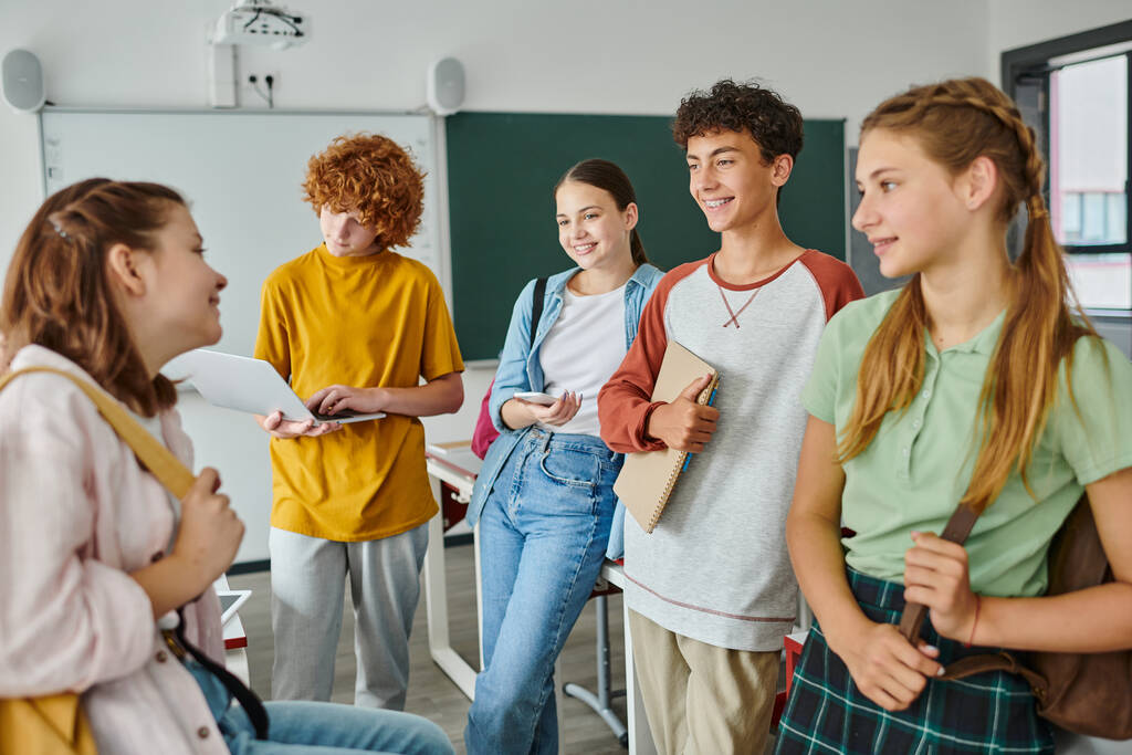 Pozytywne nastoletnie dzieci z urządzeniami i plecakami rozmawiające podczas stania w klasie w szkole - Zdjęcie, obraz
