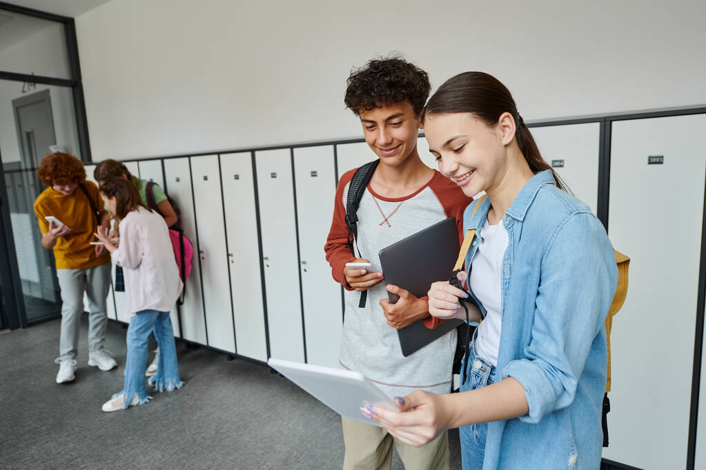 happy boy and girl looking at digital tablet and holding devices in school hallway, teen classmates - Photo, Image