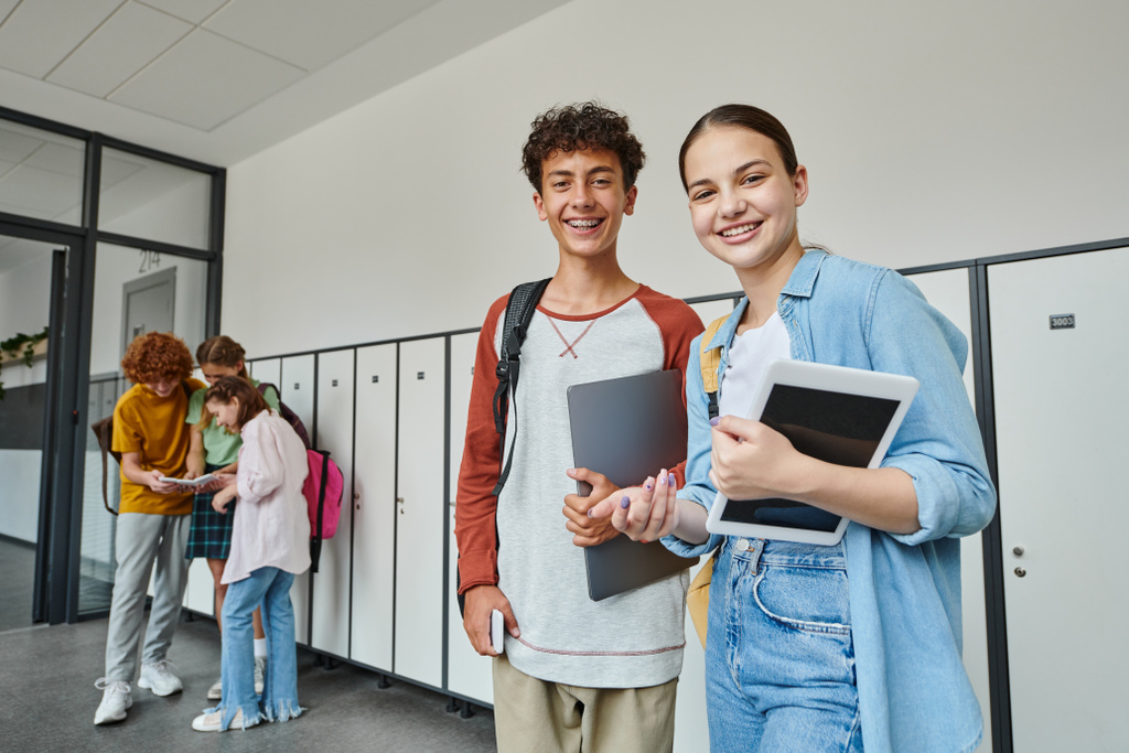 szczęśliwy nastolatek koledzy trzymając urządzenia i patrząc w kamerę w szkolnym korytarzu, młodzież studentów - Zdjęcie, obraz