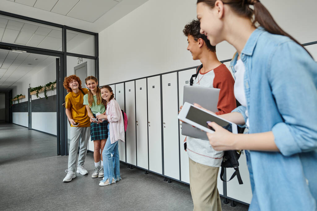estudiantes felices mirando a compañeros de clase adolescentes con dispositivos en el pasillo de la escuela, de vuelta al concepto de la escuela - Foto, imagen