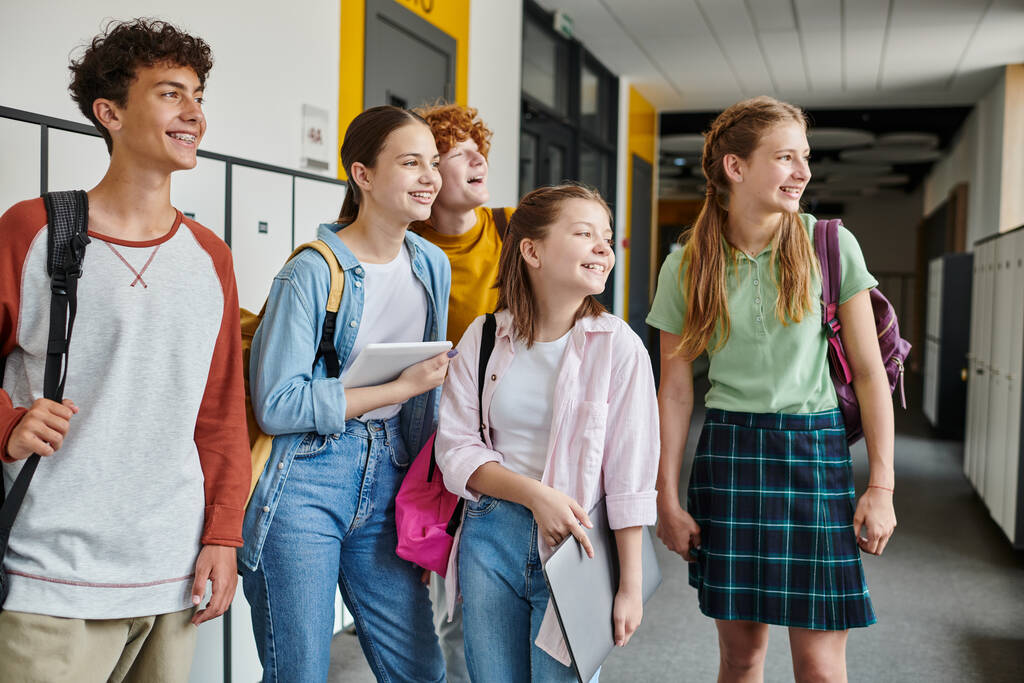 happy teenage schoolkids looking away and standing together in school hallway, teen classmates - Photo, Image
