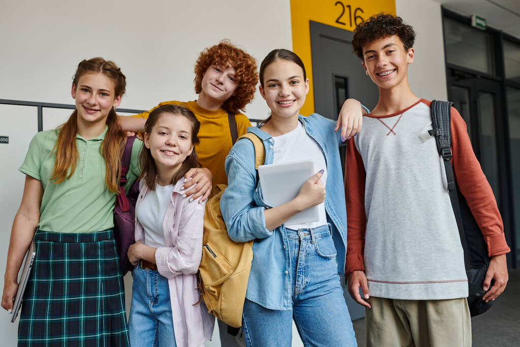повернувшись до школи, щасливі підлітки дивляться на камеру в коридорі, тримають пристрої та посміхаються разом - Фото, зображення