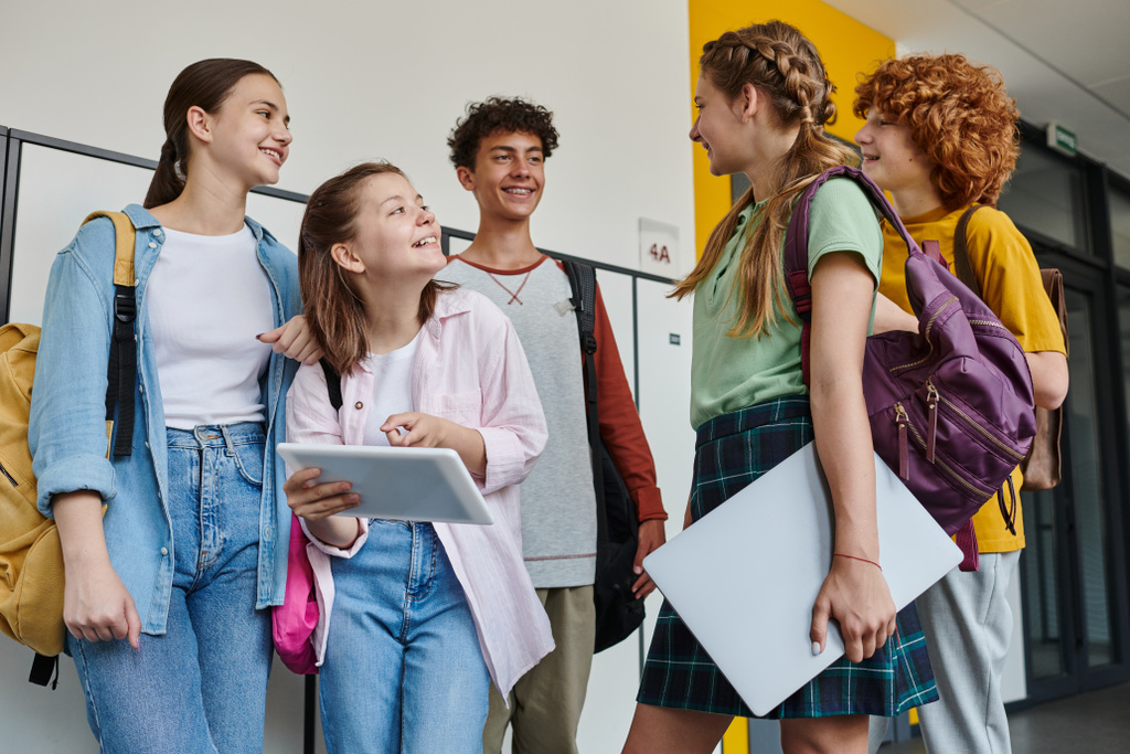 Genç kız dijital tableti işaret ediyor ve sınıf arkadaşlarına bakıyor, okul koridorundaki genç öğrenciler. - Fotoğraf, Görsel