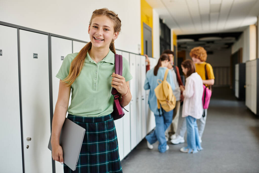 alegre adolescente sosteniendo el ordenador portátil y mirando hacia otro lado en el pasillo, borroso, escolares en el fondo - Foto, Imagen