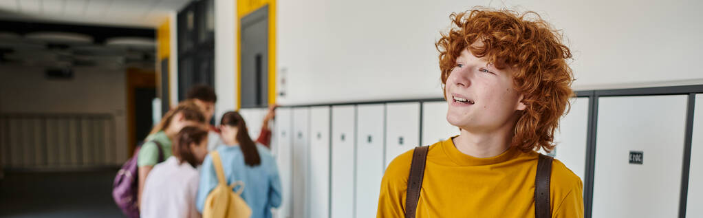 банер, рудий і кучерявий школяр дивиться геть, посміхаючись у шкільному коридорі, розмитий, студенти - Фото, зображення