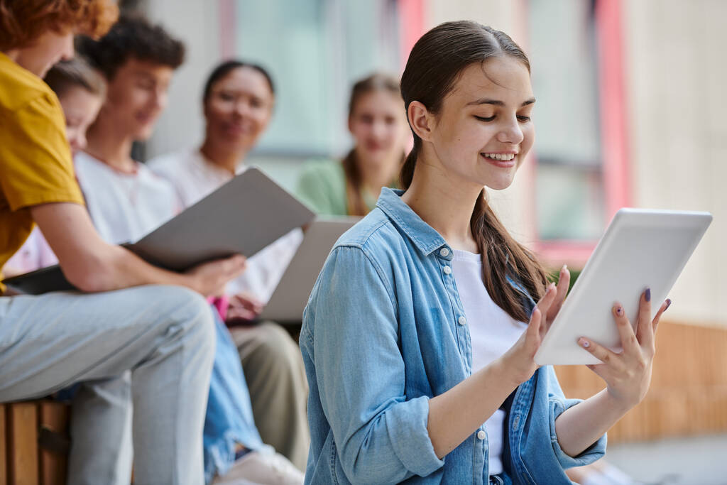 счастливая девочка-подросток улыбается и с помощью цифрового планшета рядом размытые одноклассники и учитель, разнообразие - Фото, изображение