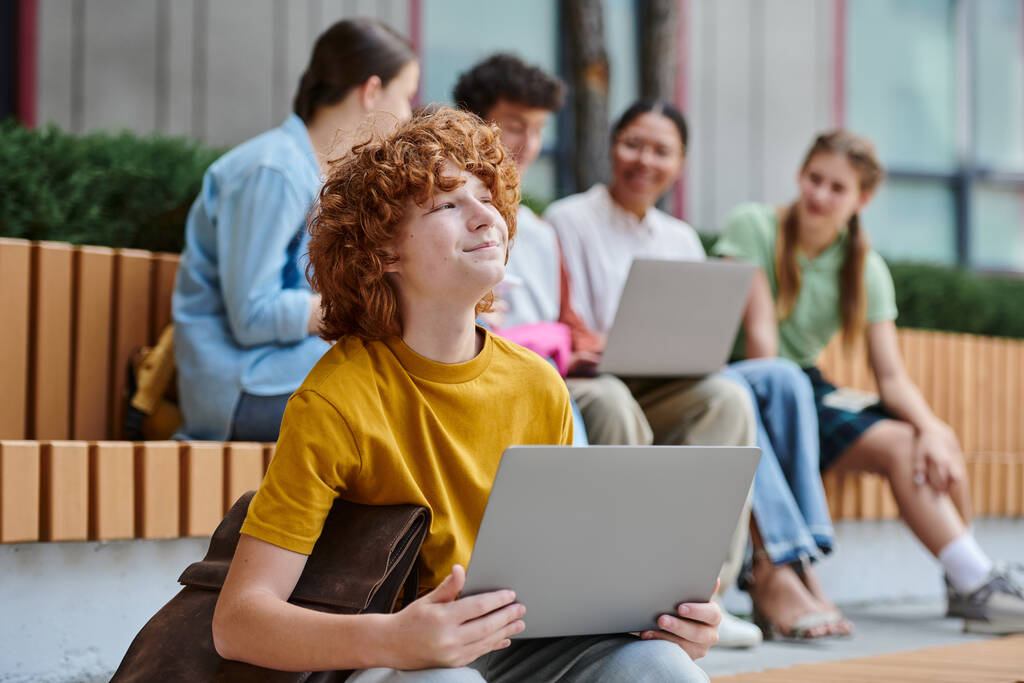chico pelirrojo de ensueño con el pelo rojo rizado sonriendo y sosteniendo el ordenador portátil, romper, desenfoque, diversidad, estudiantes - Foto, imagen