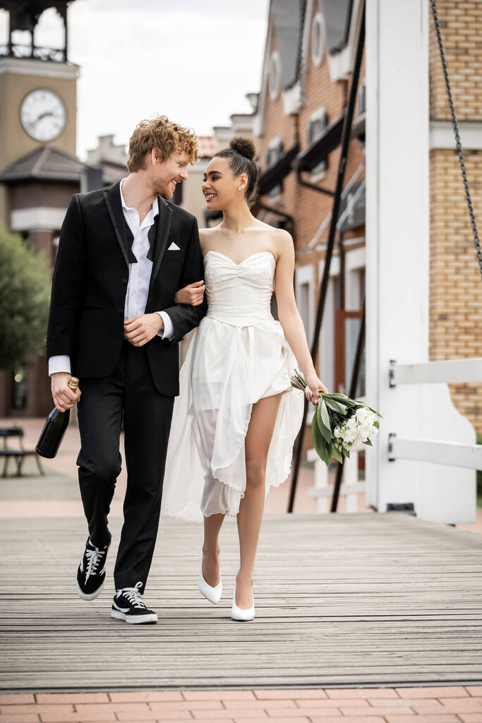 γάμος σε εξωτερικούς χώρους, πολυεθνικοί νεόνυμφοι με σαμπάνια και λουλούδια περπατώντας στη γέφυρα της πόλης - Φωτογραφία, εικόνα