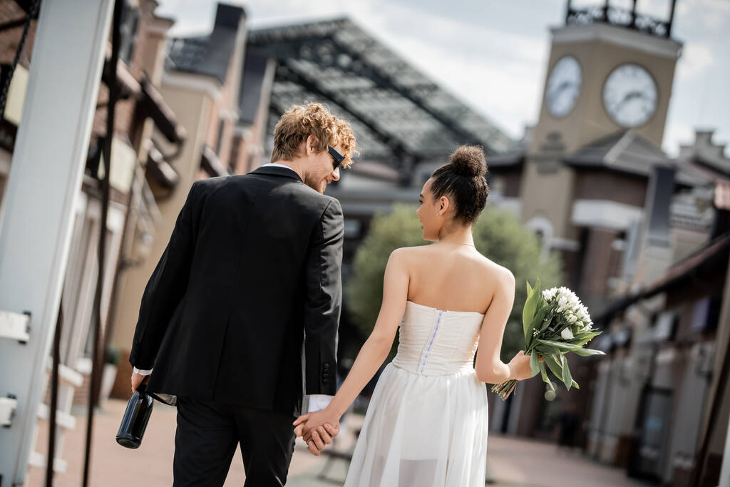 Rückansicht eines eleganten gemischtrassigen Paares mit Champagner und Blumen in der Stadt, Hochzeit im Freien - Foto, Bild