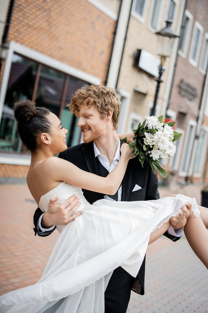 stedelijke romantiek, roodharige bruidegom met Afrikaanse Amerikaanse bruid met bruiloft boeket op straat - Foto, afbeelding