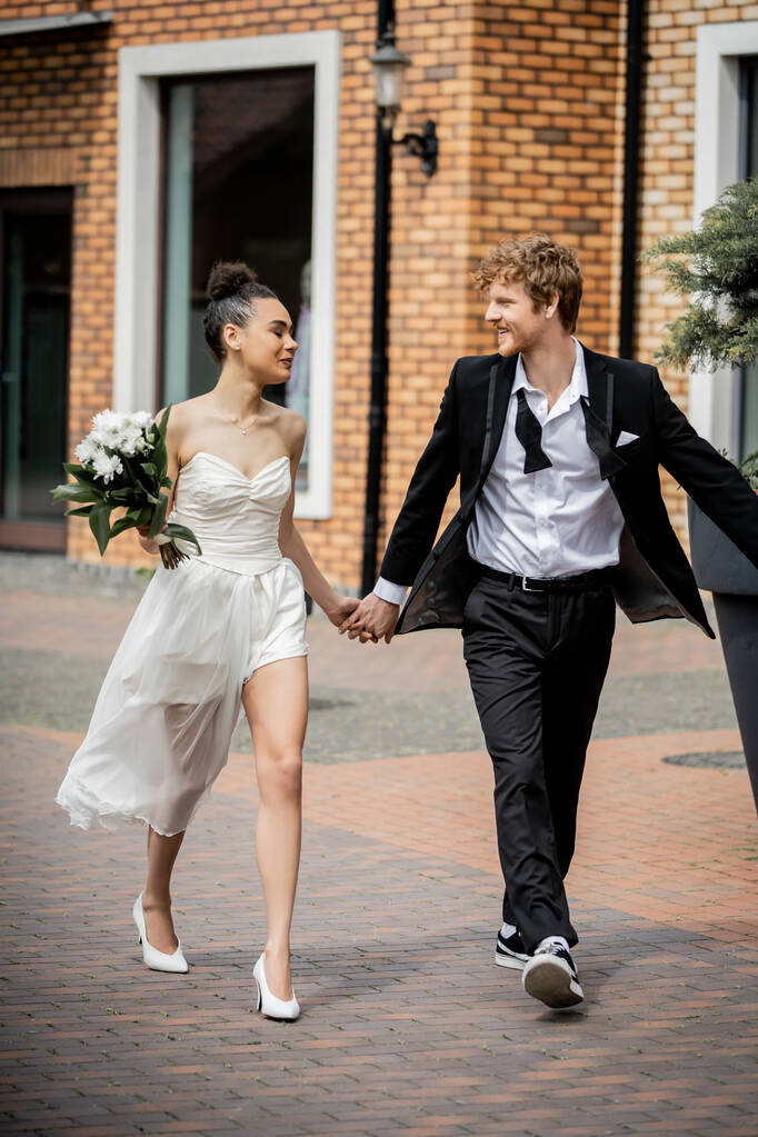african american bride with flowers and redhead groom in suit walking in city, love outdoors - Photo, Image