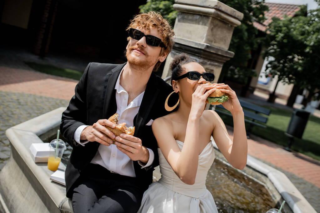 novia afroamericana comiendo hamburguesa cerca del novio pelirrojo en gafas de sol cerca de la fuente, boda al aire libre - Foto, Imagen