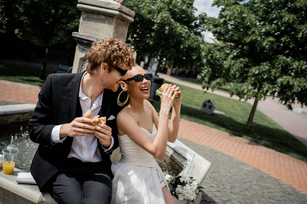 izgatott multietnikus pár ünnepli esküvő közelében városi szökőkút, napszemüveg, hamburger, narancslé - Fotó, kép