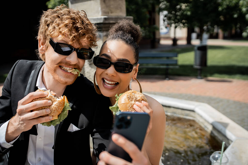 interracial newlyweds in sunglasses holding burgers and taking selfie on smartphone in city - Photo, Image