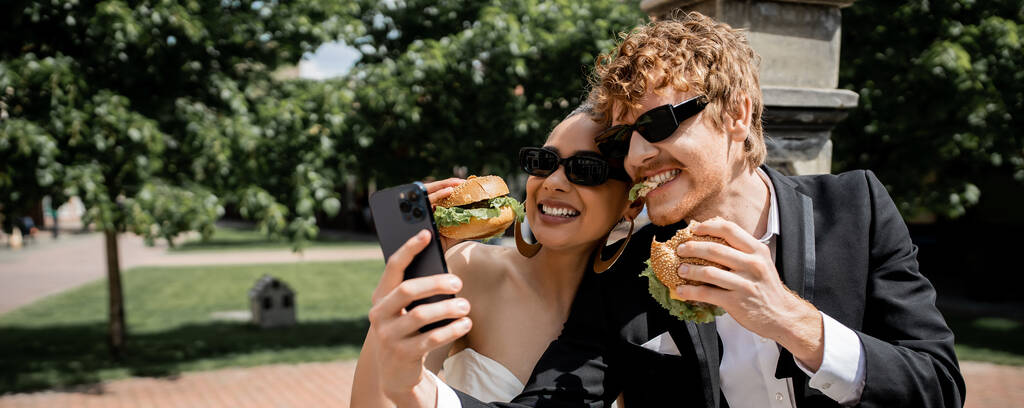 ハンバーガーを食べてスマートフォンでセルフィーを取るサングラスの異人種間の新しいweds,バナー - 写真・画像