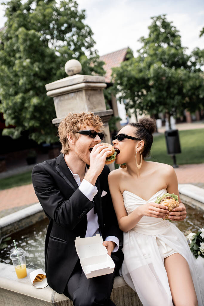 couple interracial en tenue de mariage et lunettes de soleil manger hamburger ensemble près de la fontaine en ville - Photo, image