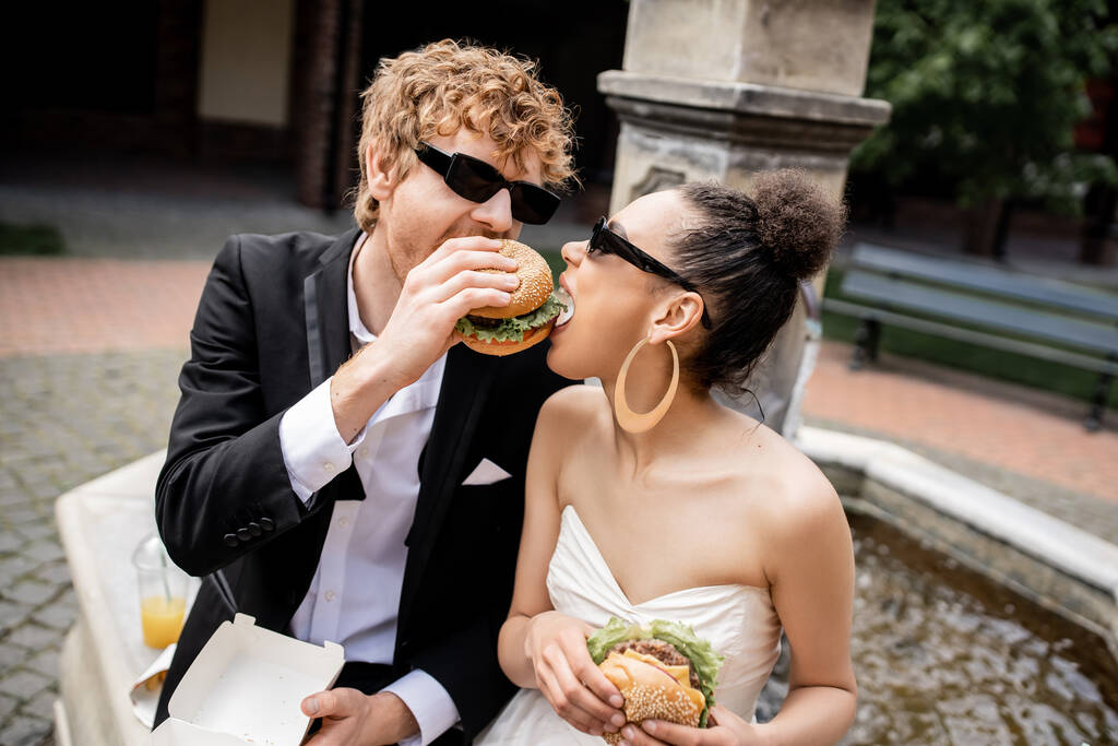 міжрасові молодята в сонцезахисних окулярах кусають весело і кусають бургер разом біля фонтану - Фото, зображення