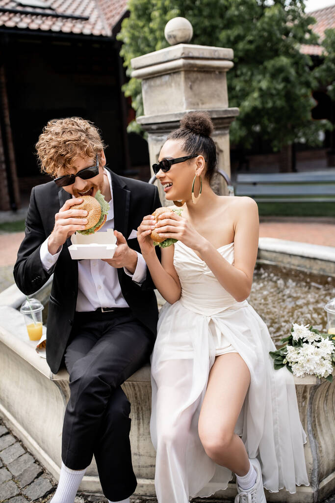 рудий чоловік в сонцезахисних окулярах їсть бургер біля афроамериканської нареченої, сидить на міському фонтані - Фото, зображення