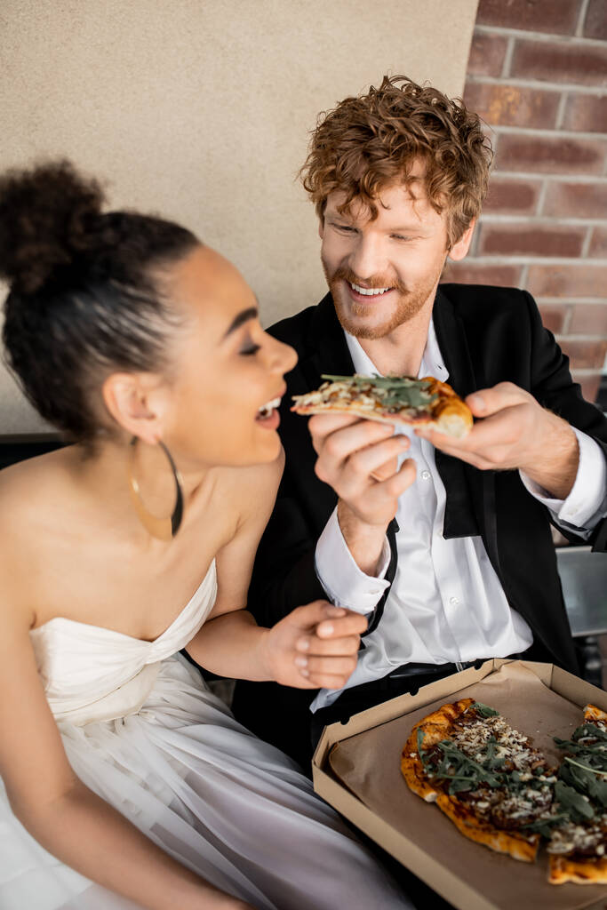 redhead groom feeding overjoyed bride with tasty pizza on bench, outdoor wedding in european city - Photo, Image