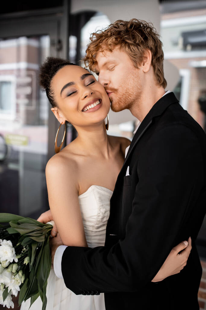 rothaariger Bräutigam im schwarzen Anzug umarmt und küsst stylische afrikanisch-amerikanische Braut auf der Stadtstraße - Foto, Bild
