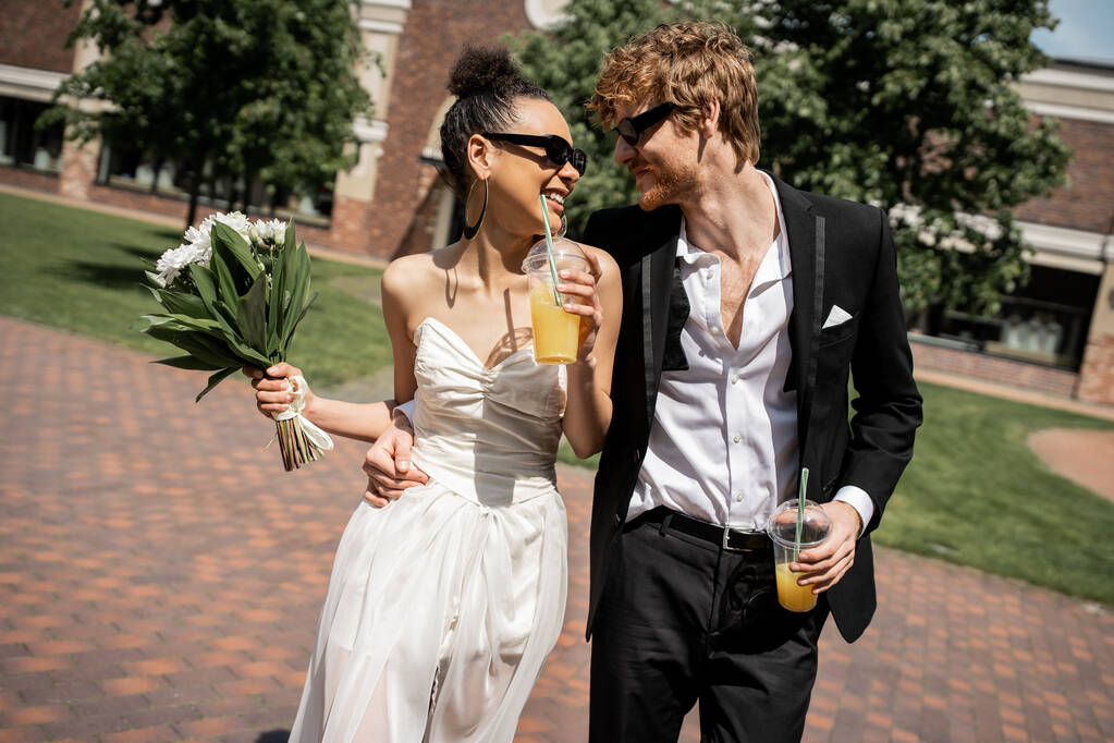 fröhliches multiethnisches Brautpaar in Sonnenbrille, das mit Orangensaft spaziert, Hochzeit auf der städtischen Straße - Foto, Bild