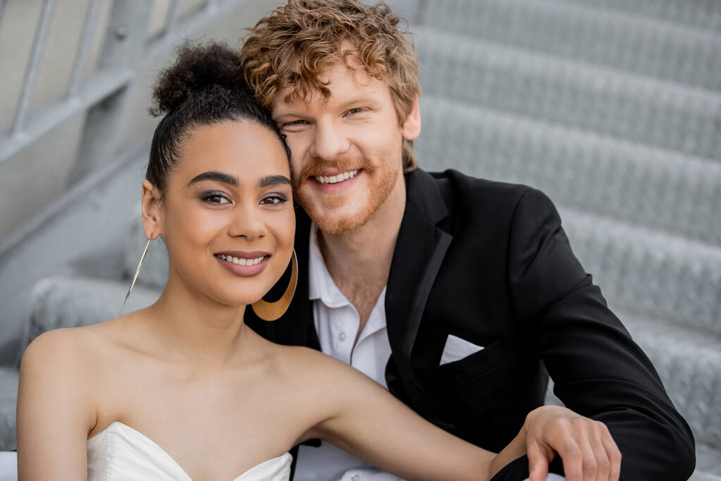 portrait of carefree and stylish interracial newlyweds smiling at camera, outdoor wedding - Photo, Image