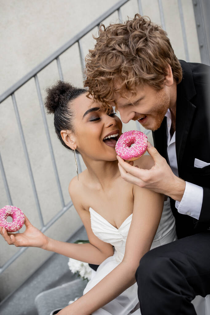 cheerful multiethnic newlyweds biting sweet donut, wedding celebration on city street - Photo, Image